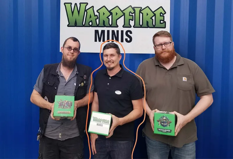 The three Warpfire Minis founders holding Warhammer boxes under their shop sign